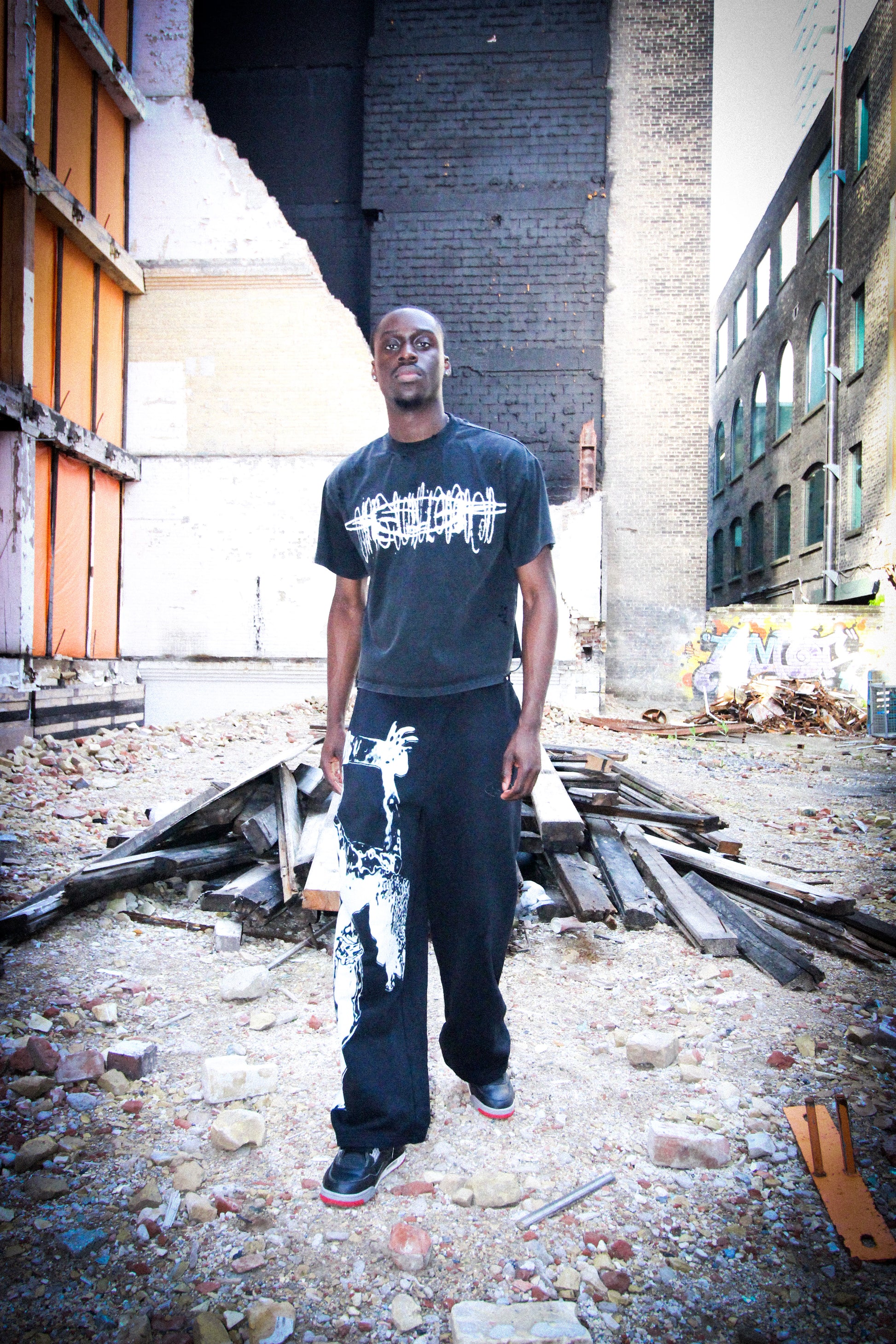 Man standing in an urban decay setting wearing a dark t-shirt with white graphics and dark pants.
